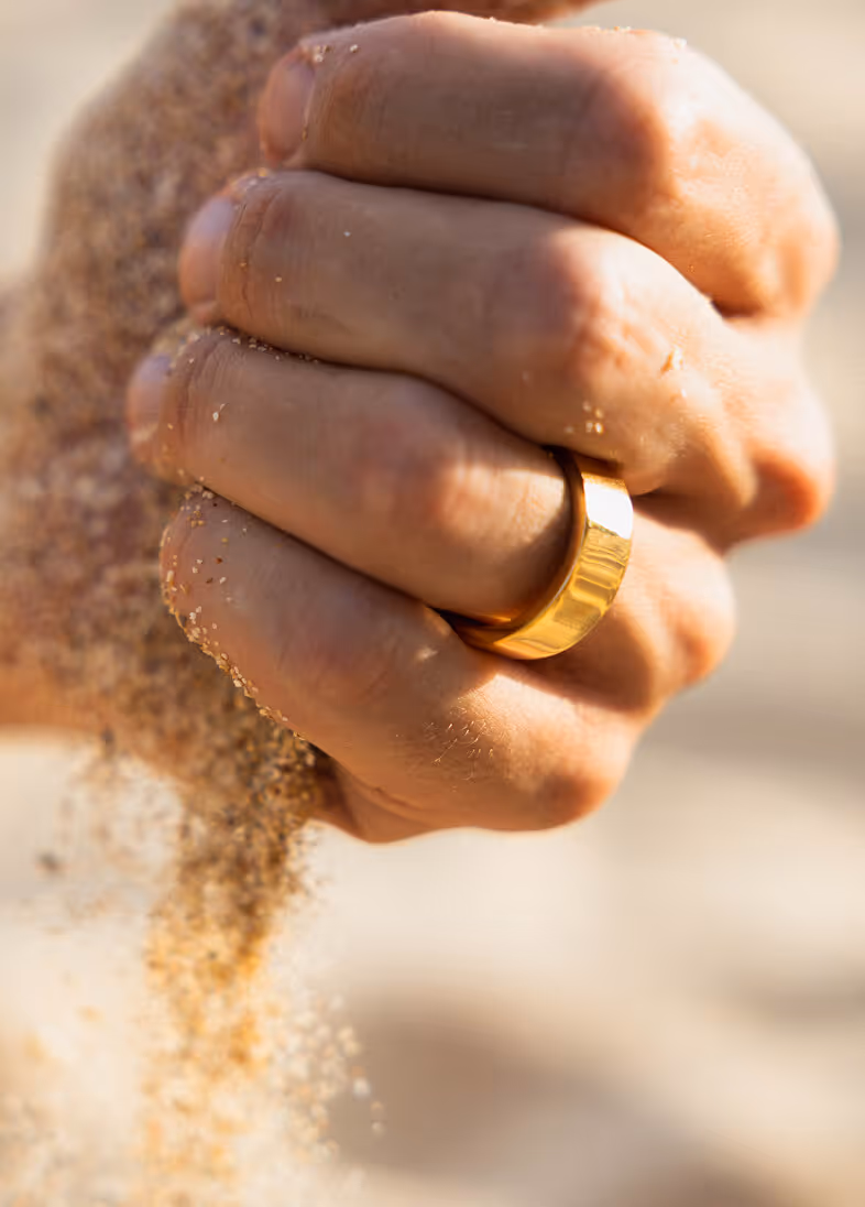 Close-up of a hand with sand falling through fingers, wearing a shiny gold ring on the finger.