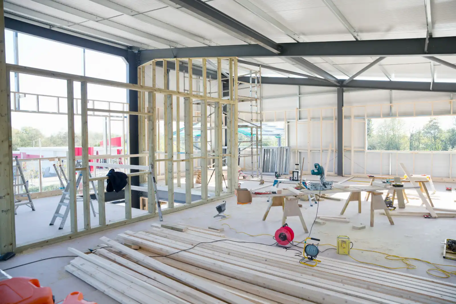Interior of a construction site with wooden framing, ladders, construction tools, and scattered lumber under a metal roof.