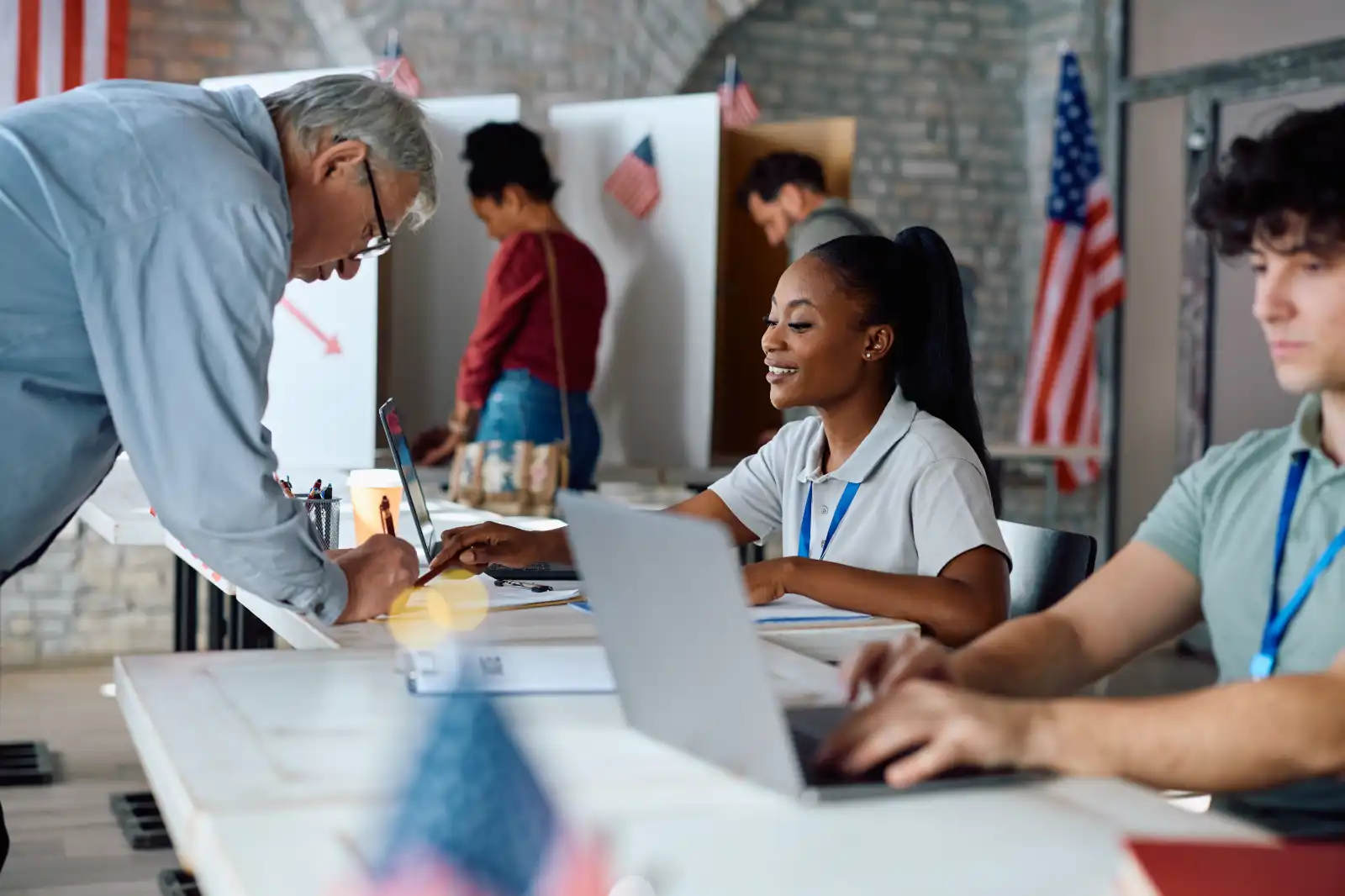 A woman voter registration official helps an elderly man sign documents at a polling station with US flags in the background.