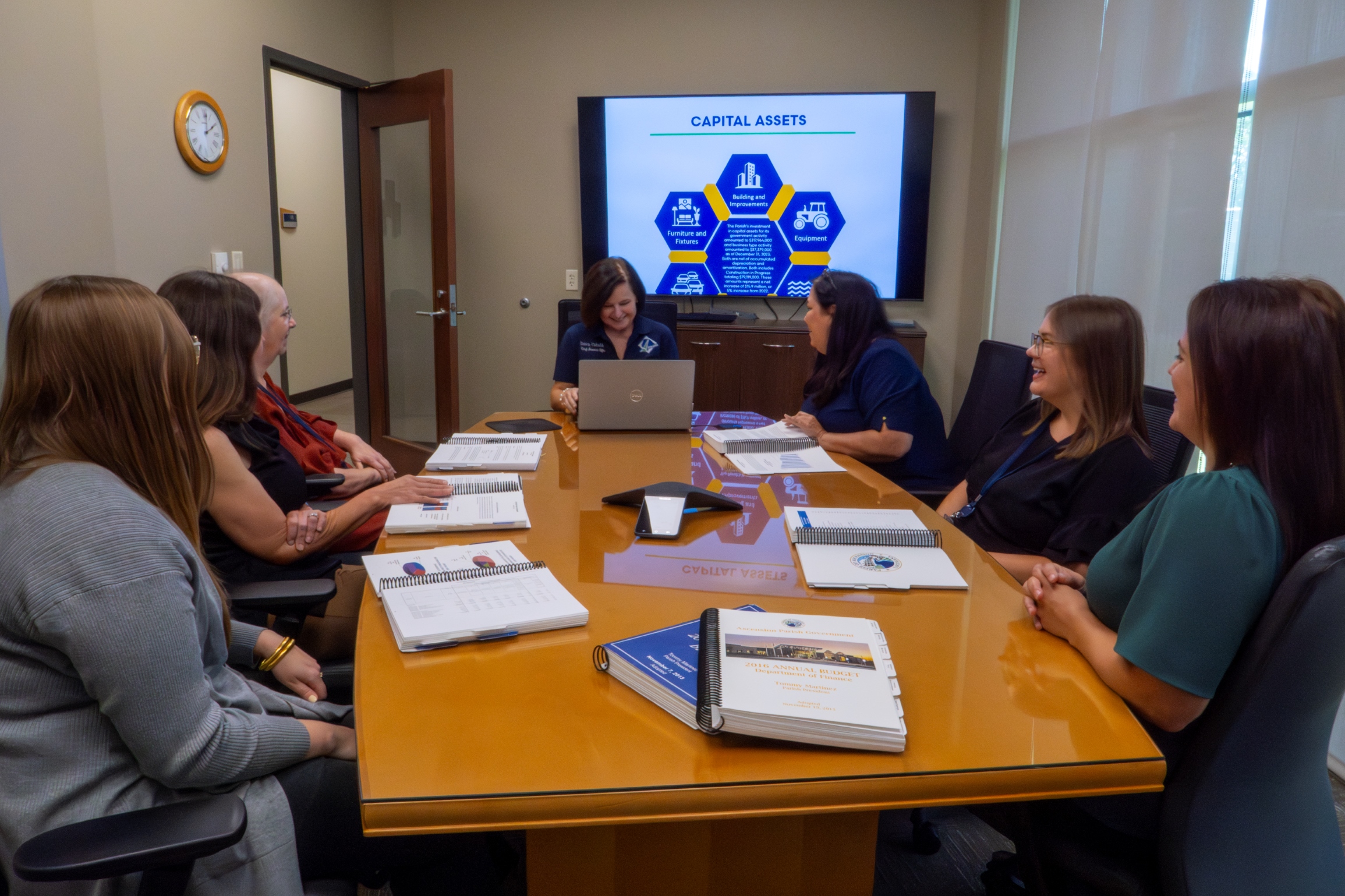 Six women in a conference room are engaged in a meeting with documents and a presentation on capital assets displayed on a screen.