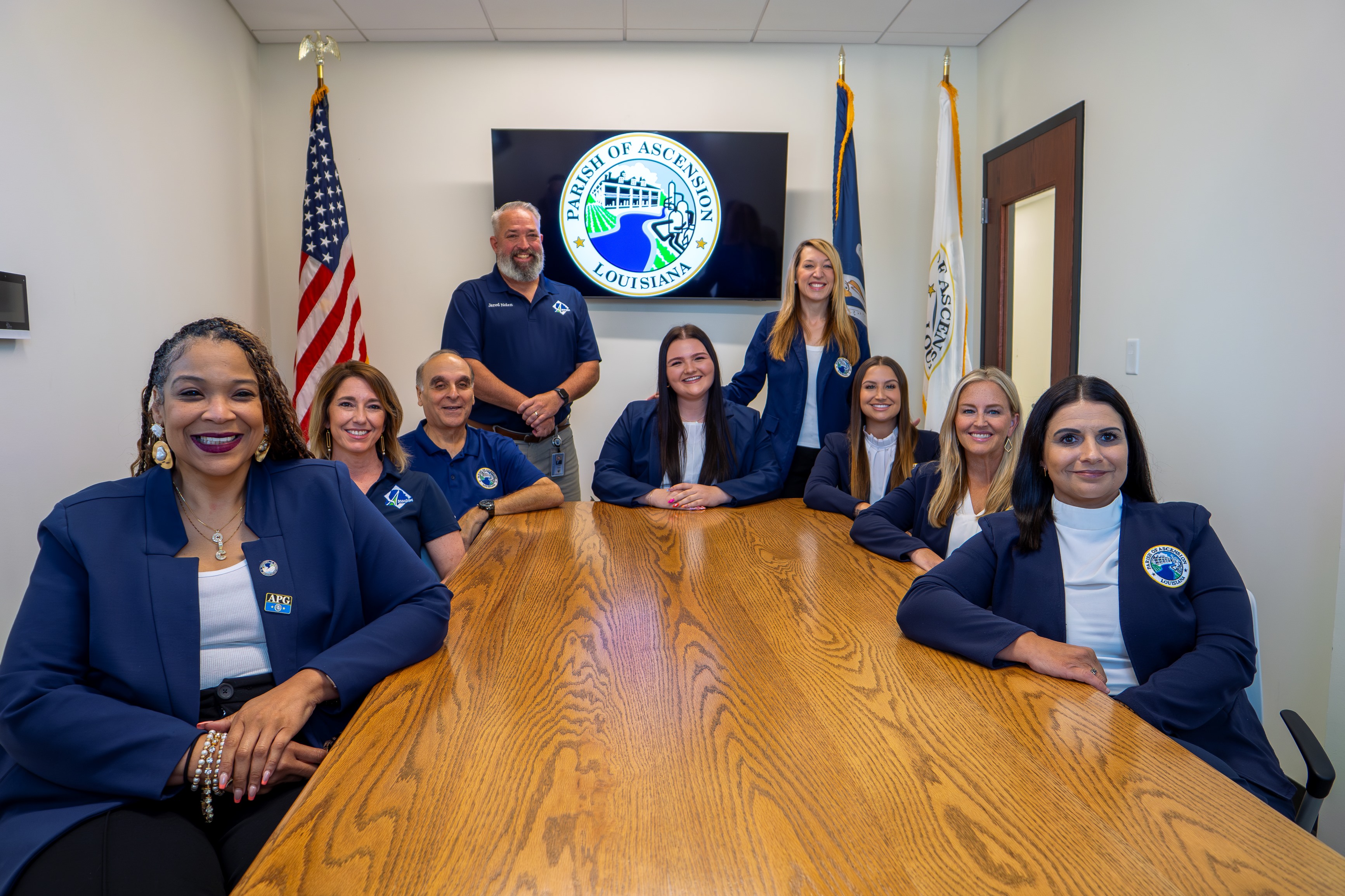 Group of eight people, five women seated and three standing, around a conference table with an American flag, Louisiana state flag, and Parish of Ascension flag behind them.