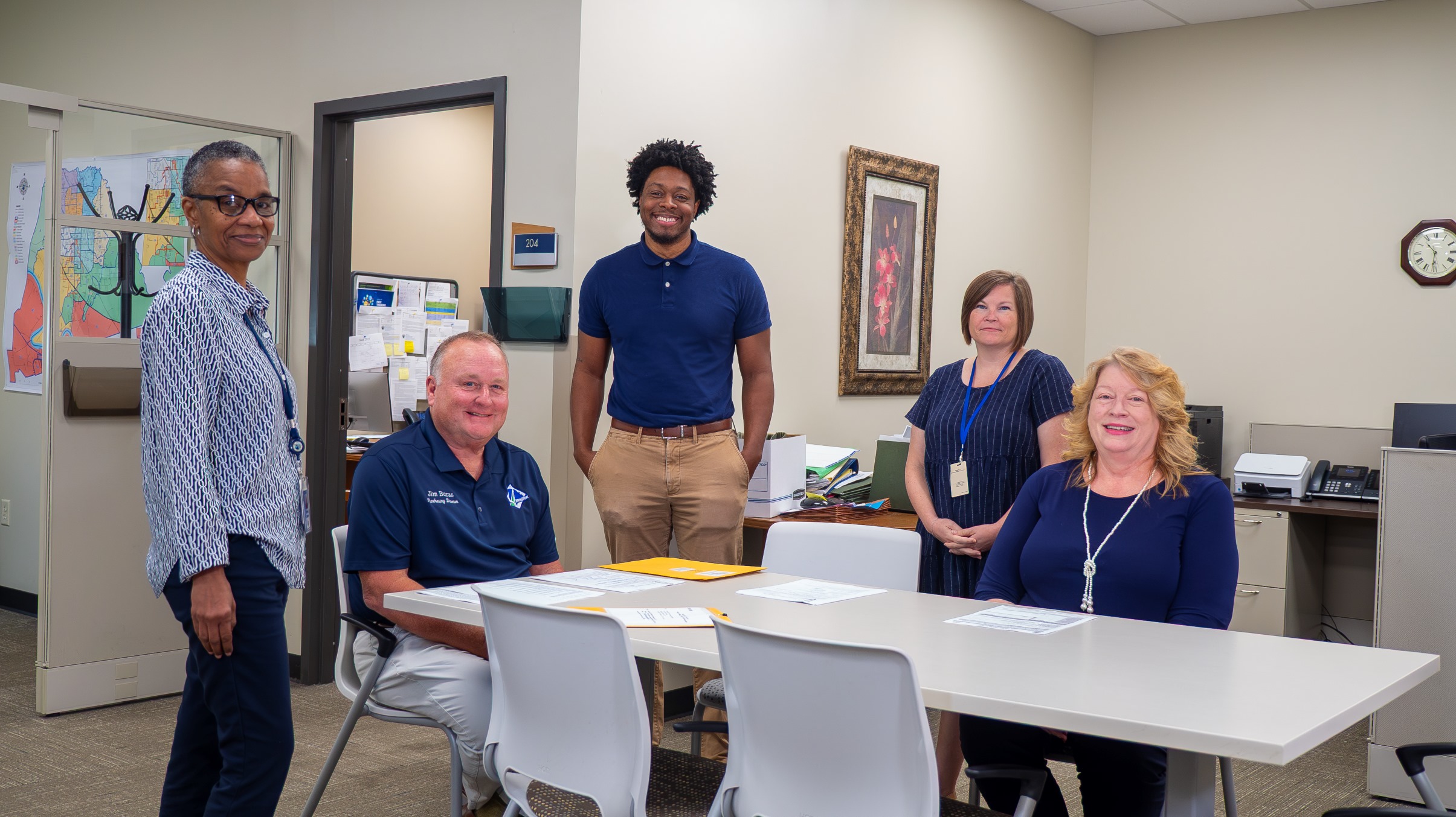 Five office workers, three women and two men, smiling in a meeting room with a table and chairs.