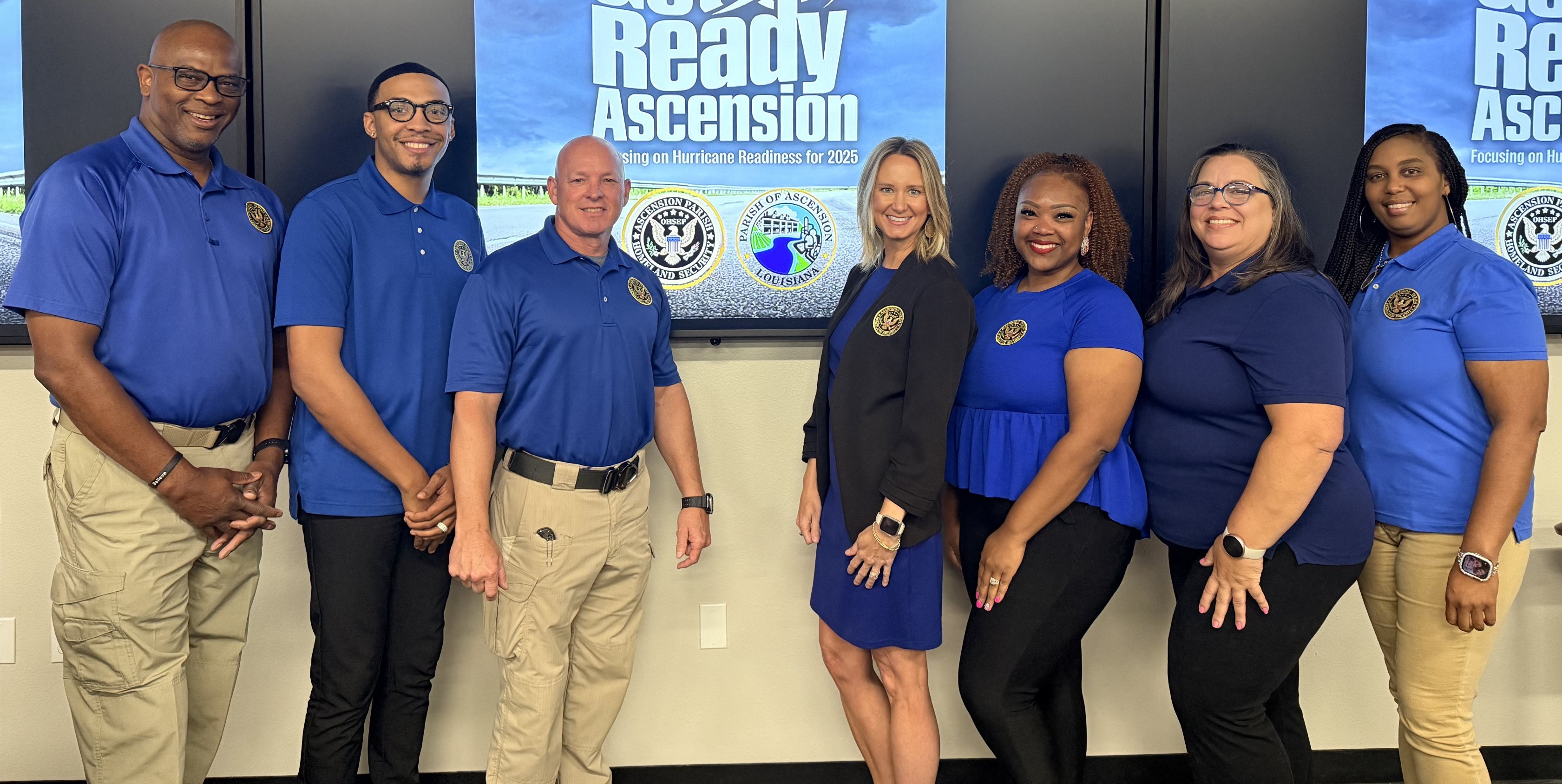 Seven people wearing blue shirts with official badges standing indoors against screens displaying 'Get Ready Ascension' hurricane readiness campaign.