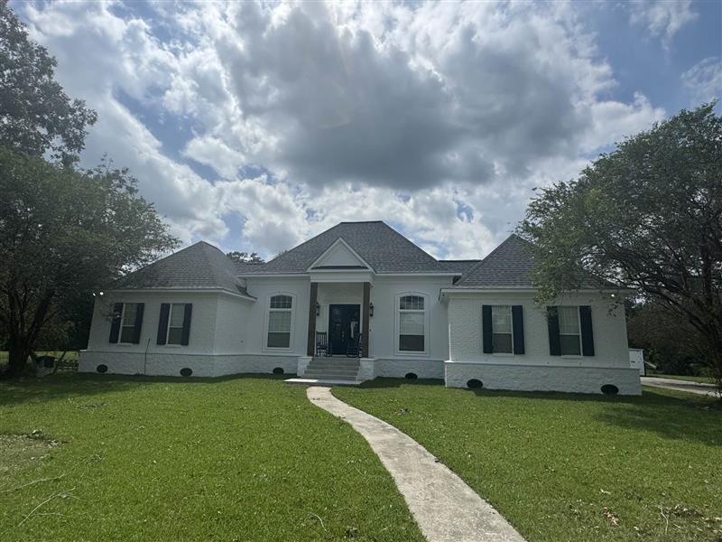 Single-story white brick house with dark roof, black shutters, and a curved concrete walkway on a grassy lawn under a cloudy sky.