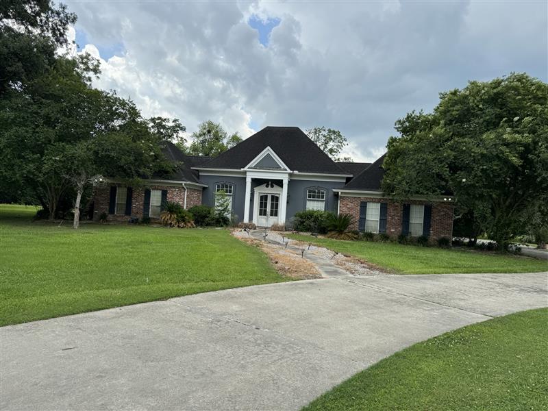 Single-story house with a dark roof, white front door with columns, and brick walls, surrounded by green lawn and trees under a cloudy sky.