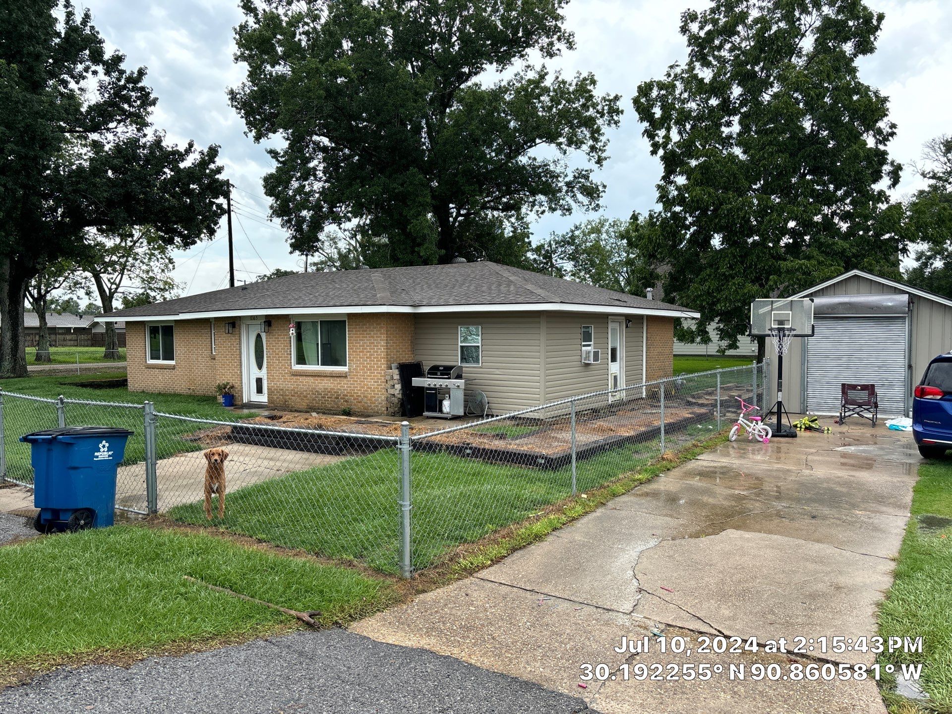 Single-story brick and siding house with a fenced front yard, a dog near the gate, a driveway with a basketball hoop, children's bike, and a blue car parked.