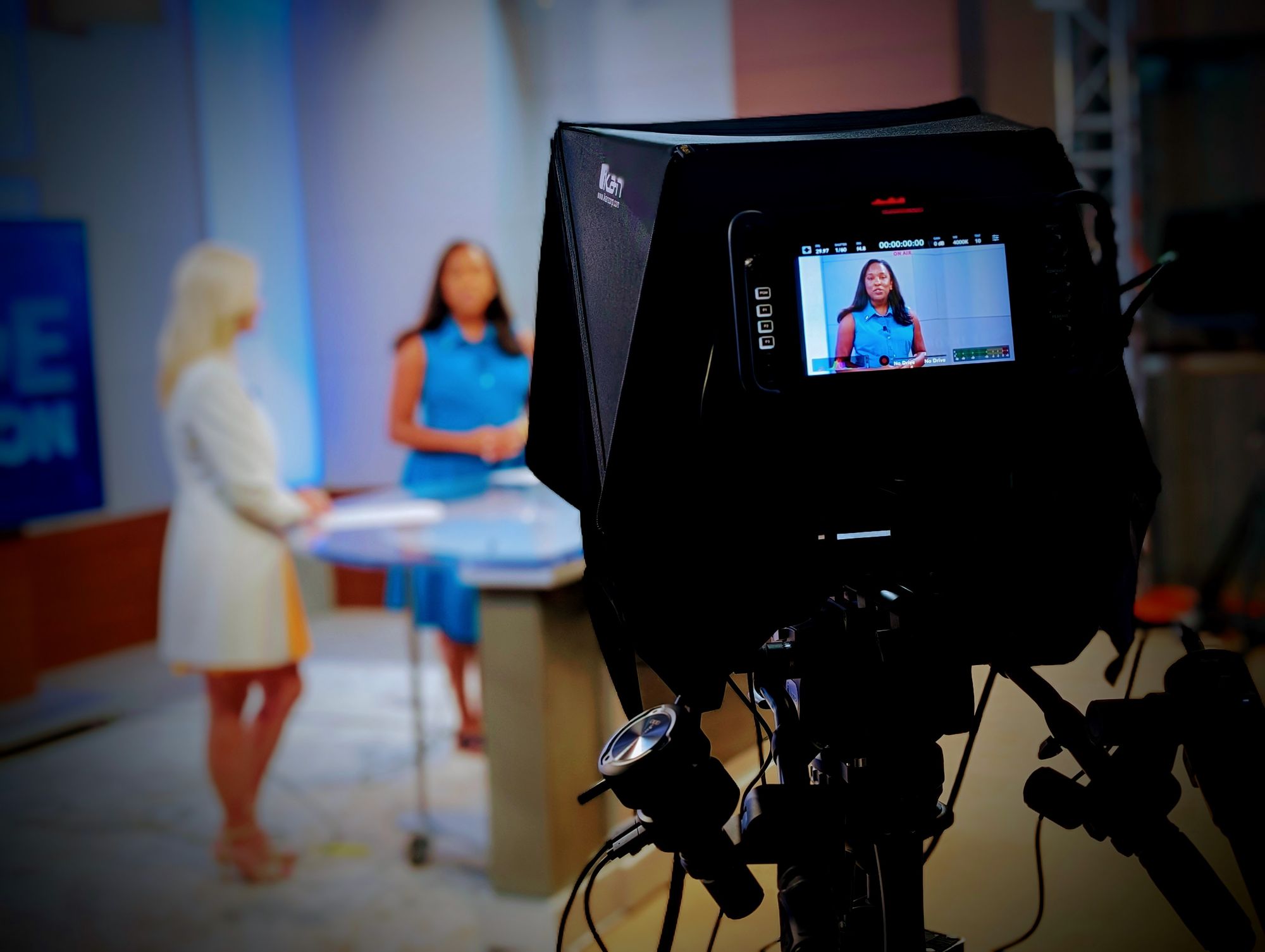 Professional video camera focusing on a woman in a blue dress standing in a TV studio with a blurred woman in a white blazer in the background.