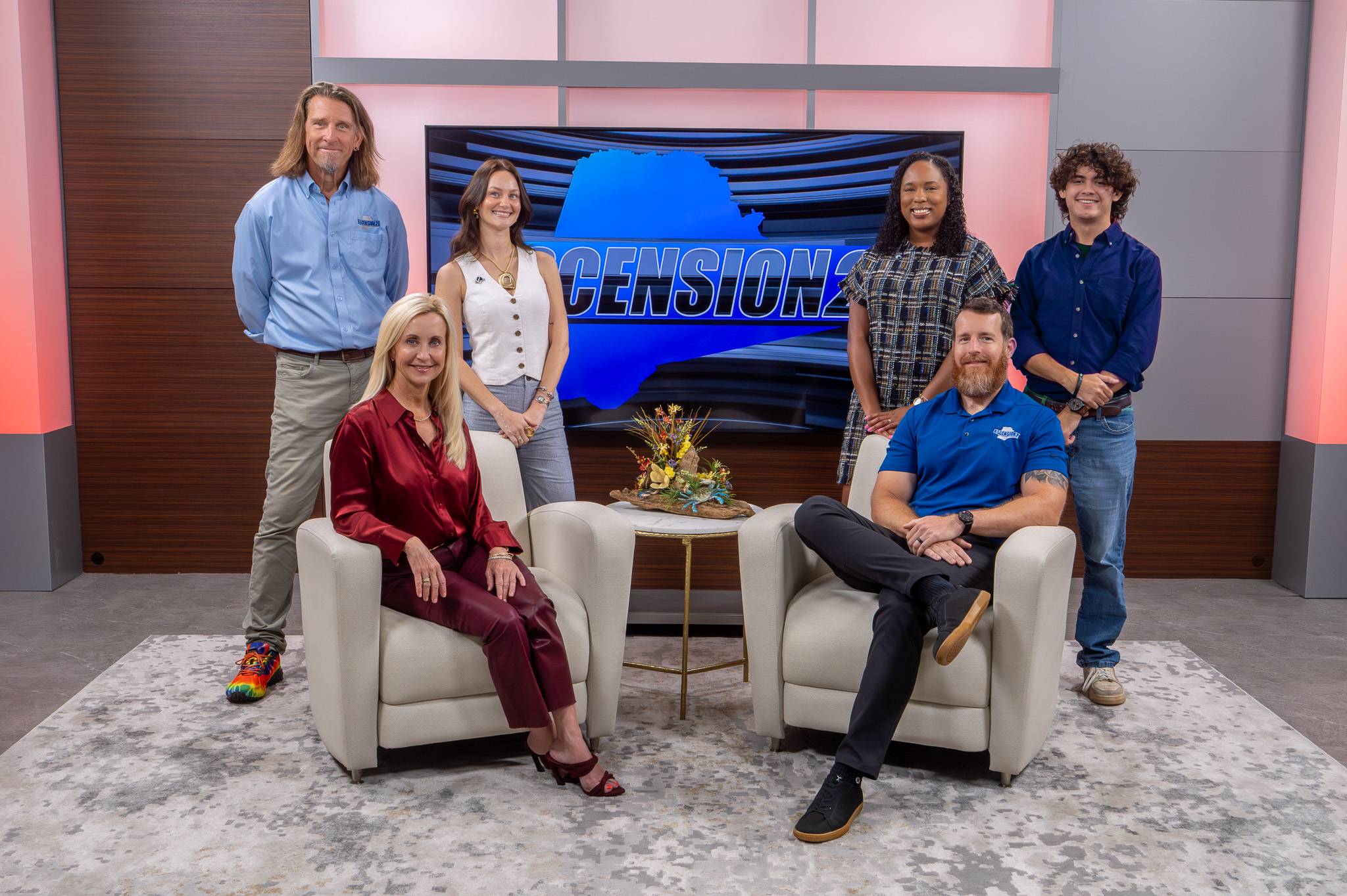 Six people posing in a studio set with two seated in armchairs and four standing behind, a TV screen behind them displays a blue graphic with the word 'ASCENSION'.