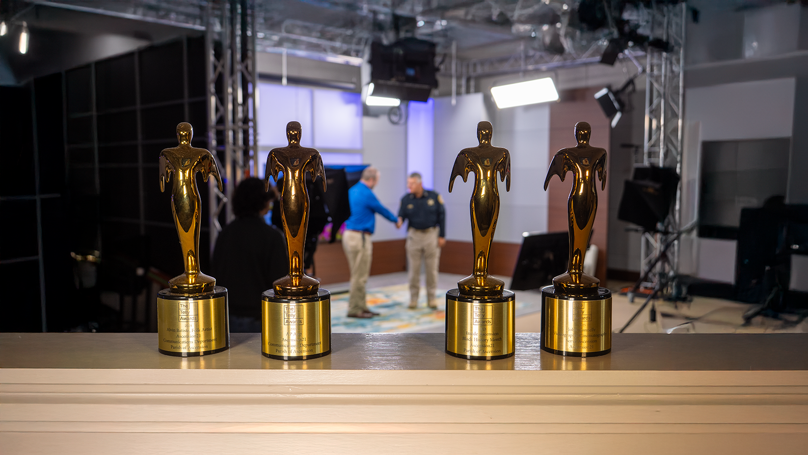 Four golden Telly Awards trophies lined up on a ledge in a studio with two men shaking hands in the blurred background.