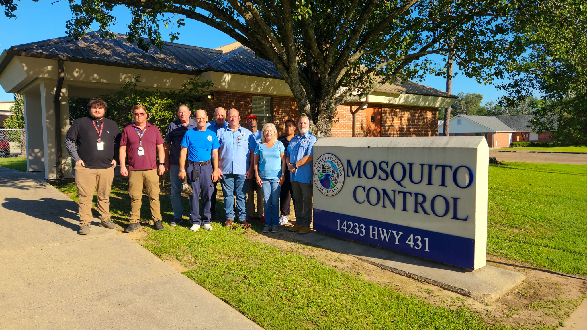 Group of nine people standing outside near a building and a sign that reads Mosquito Control 14233 HWY 431.