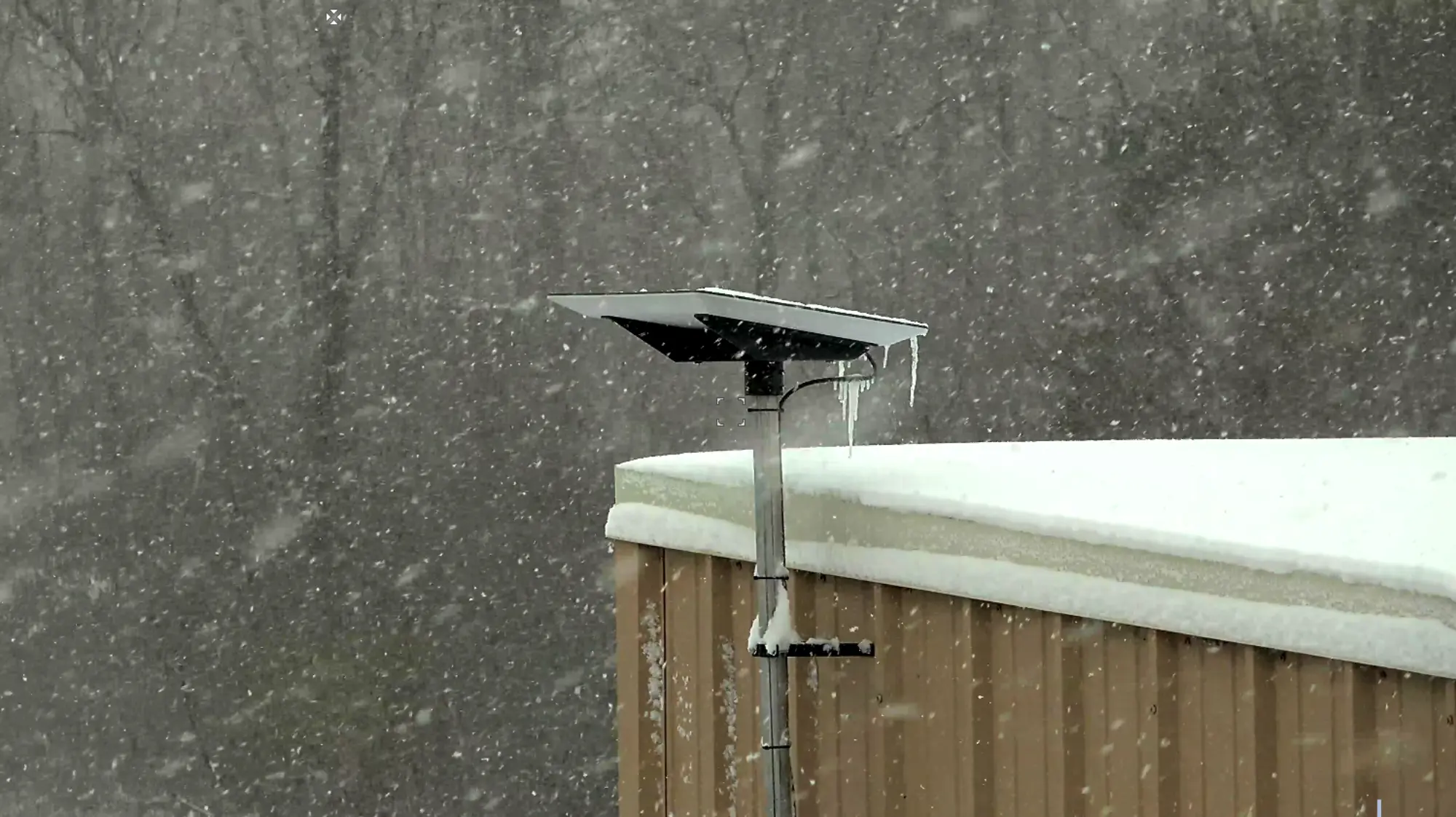 Metal weather station covered in snow and icicles mounted on a snow-covered rooftop during heavy snowfall.