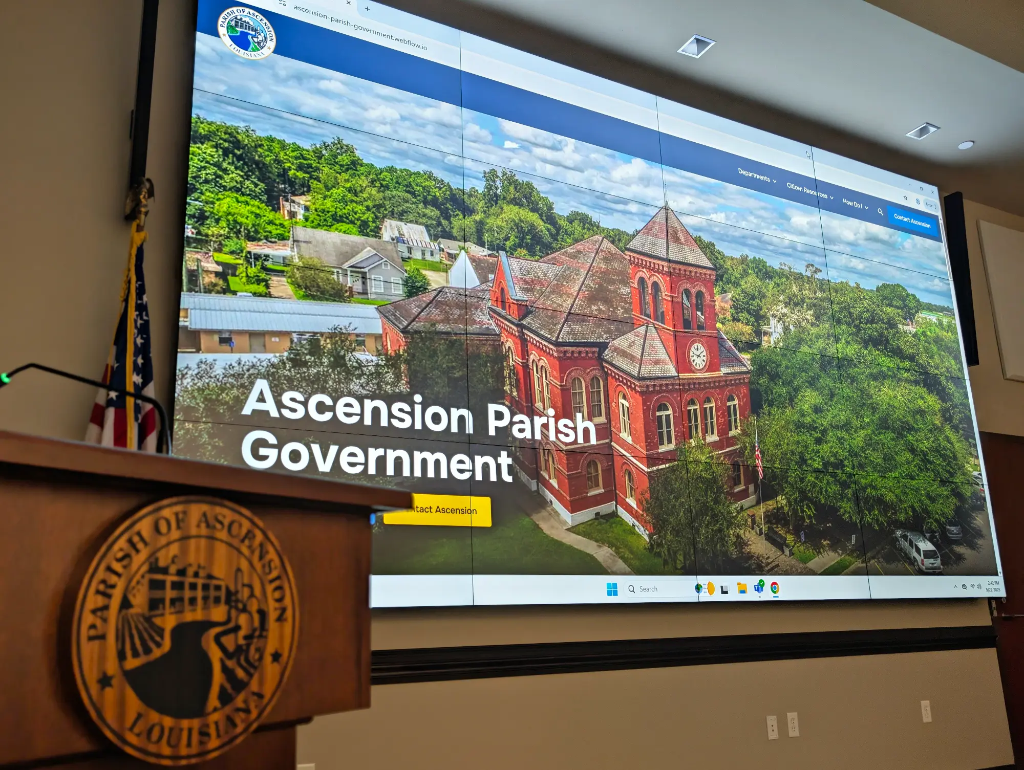 Large screen displaying the Ascension Parish Government website with an aerial view of a red brick building, with a podium in front bearing the Parish of Ascension Louisiana seal.
