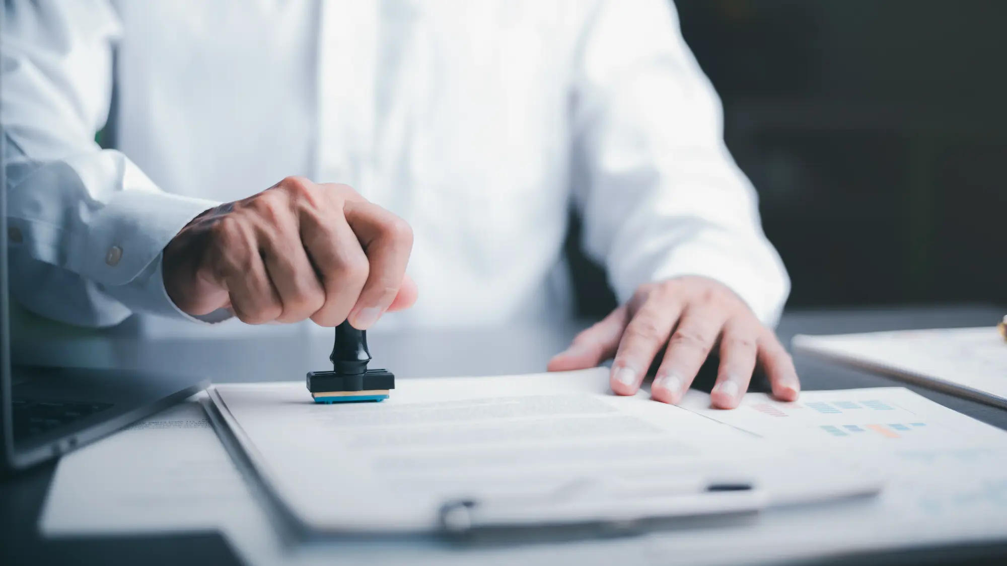 Person in a white shirt stamping a document on a desk with charts and a pen nearby.