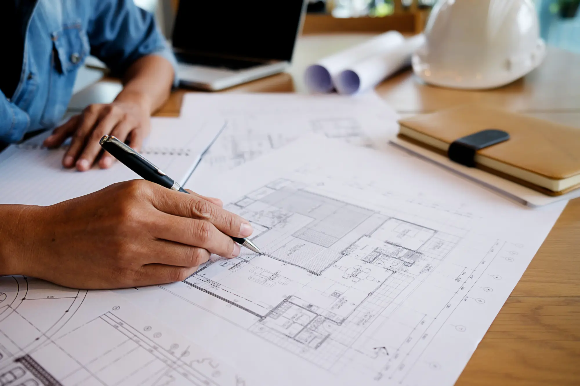 Person reviewing architectural blueprints with a pen on a desk with a laptop, notebook, and white safety helmet.