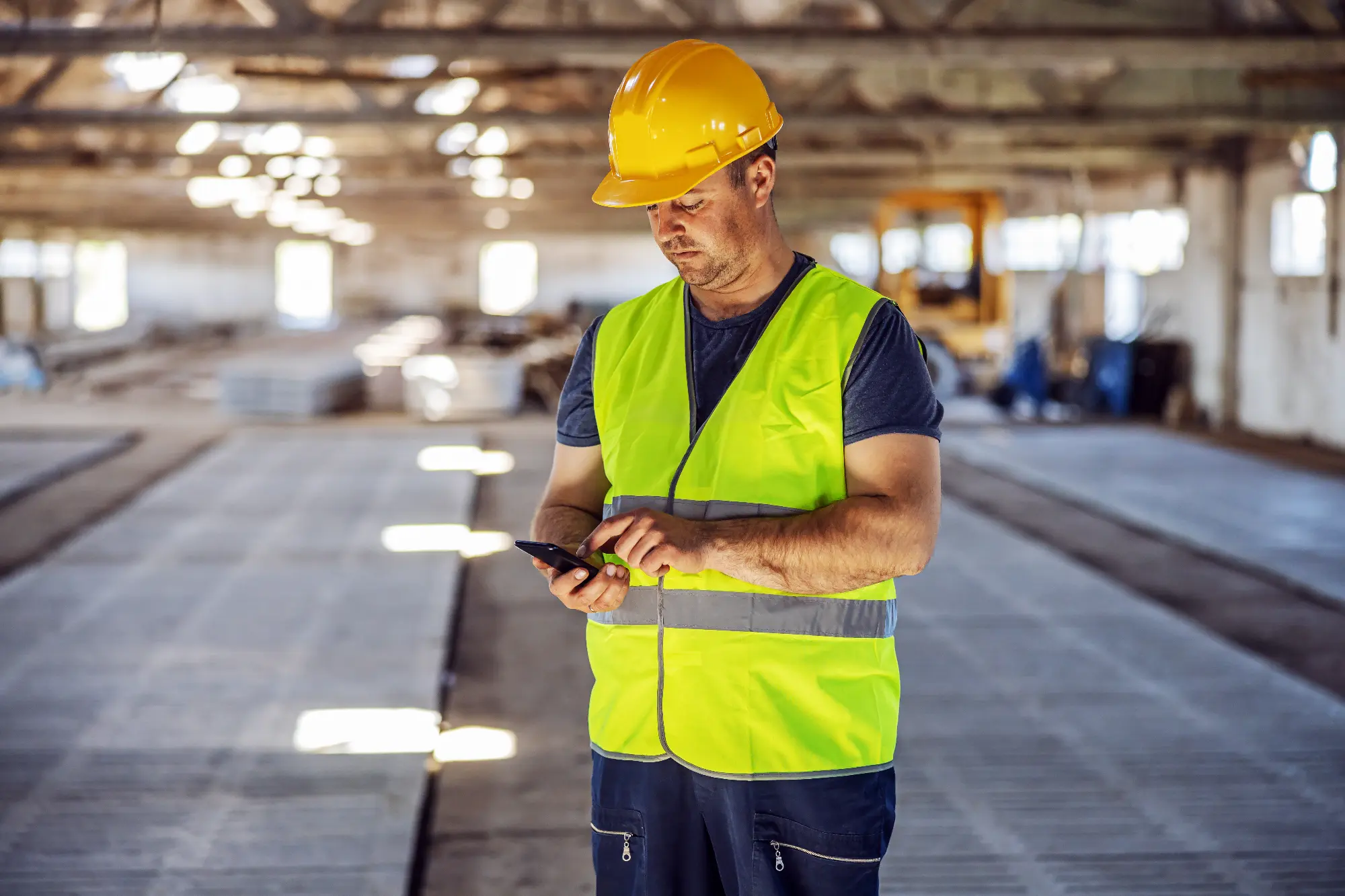 Construction worker in a yellow hard hat and reflective vest using a smartphone inside an unfinished building.