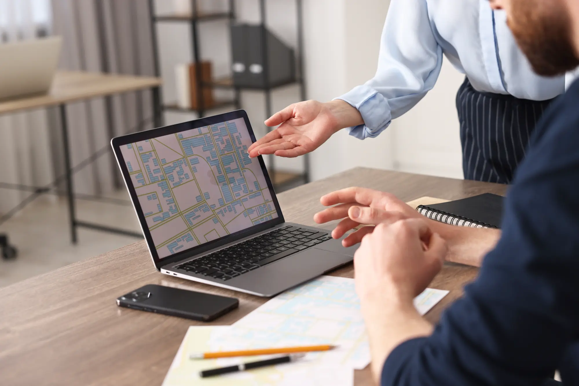 Two people discussing a digital map displayed on a laptop screen at a table with a smartphone, paper maps, and stationery.