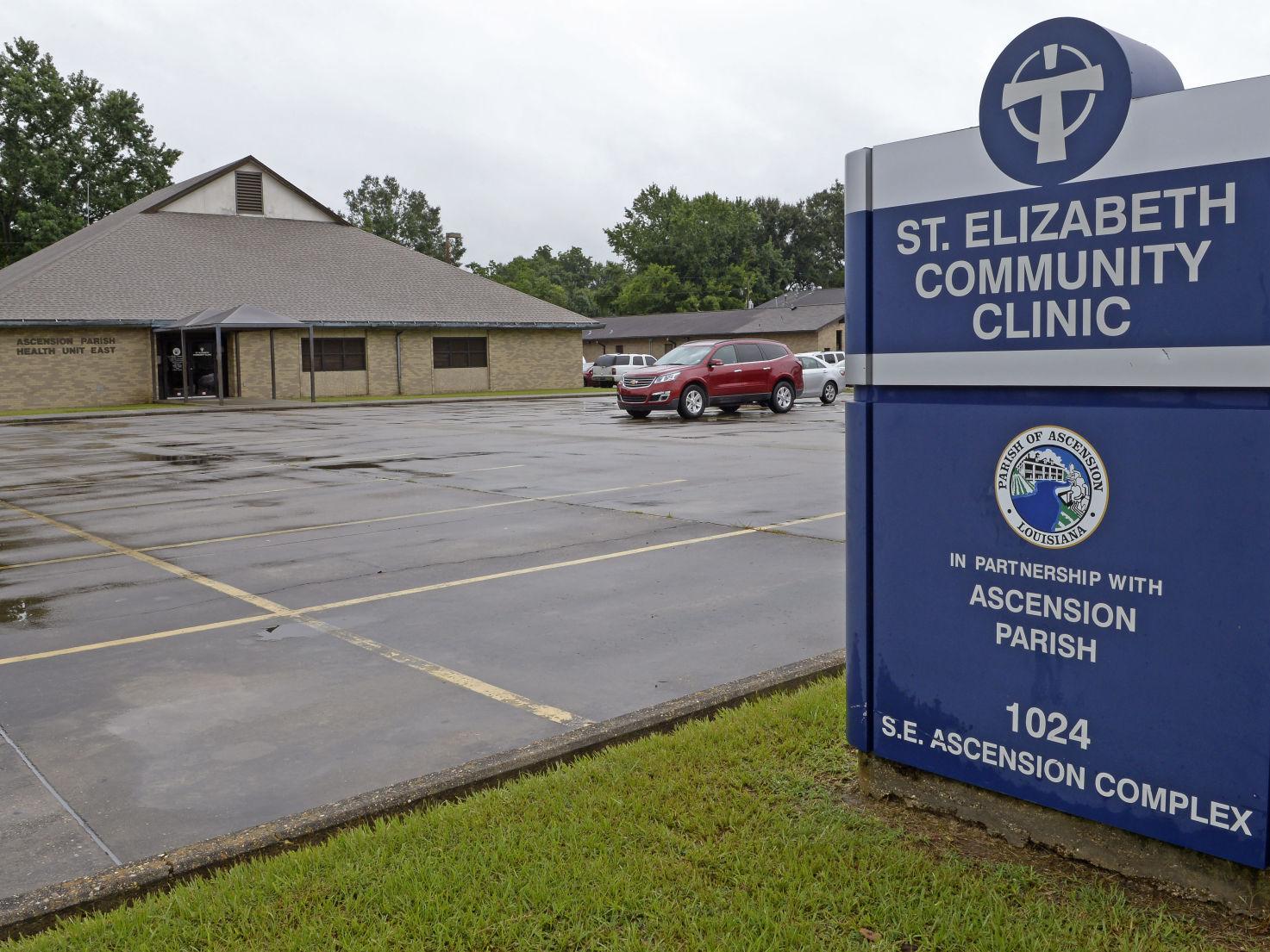 Parking lot in front of St. Elizabeth Community Clinic building with a blue sign showing its name and address in Ascension Parish, Louisiana.