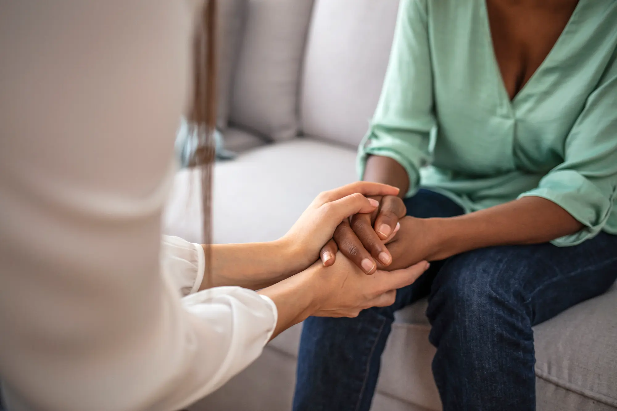 Close-up of a person in a white shirt holding the hands of another person sitting on a couch wearing a green top and jeans.