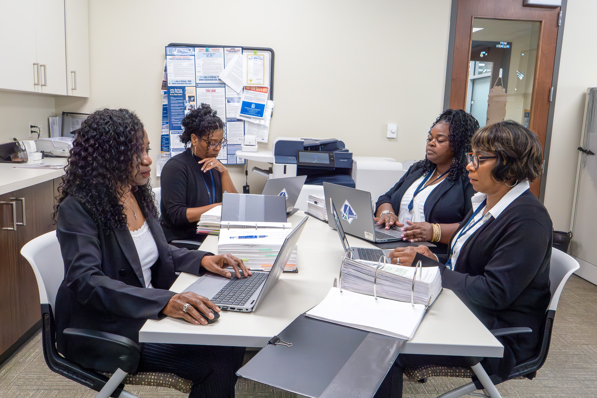 Four women seated around a table in an office working on laptops and reviewing documents.