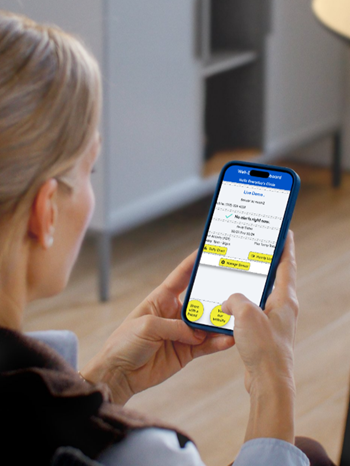 Person holding a smartphone displaying a webpage with text and yellow buttons in a room with wooden floor and gray furniture.