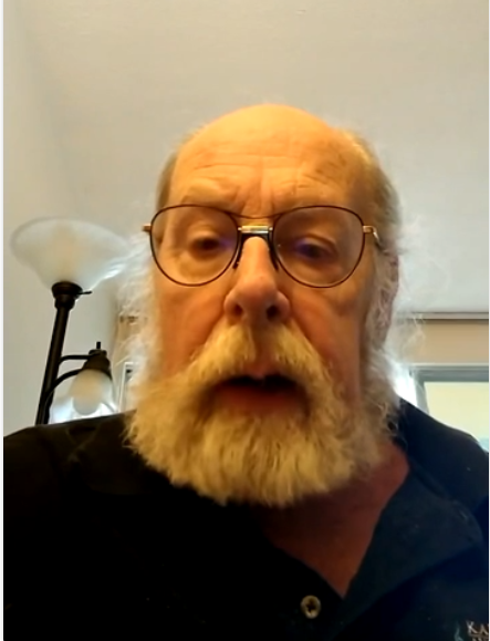Elderly man with glasses, grey beard, and mustache wearing a black polo shirt, sitting indoors near a lamp and window.