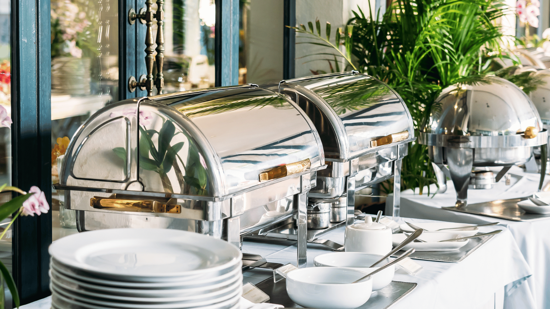 Buffet setup with stainless steel chafing dishes and white porcelain plates in an elegant catering setting.