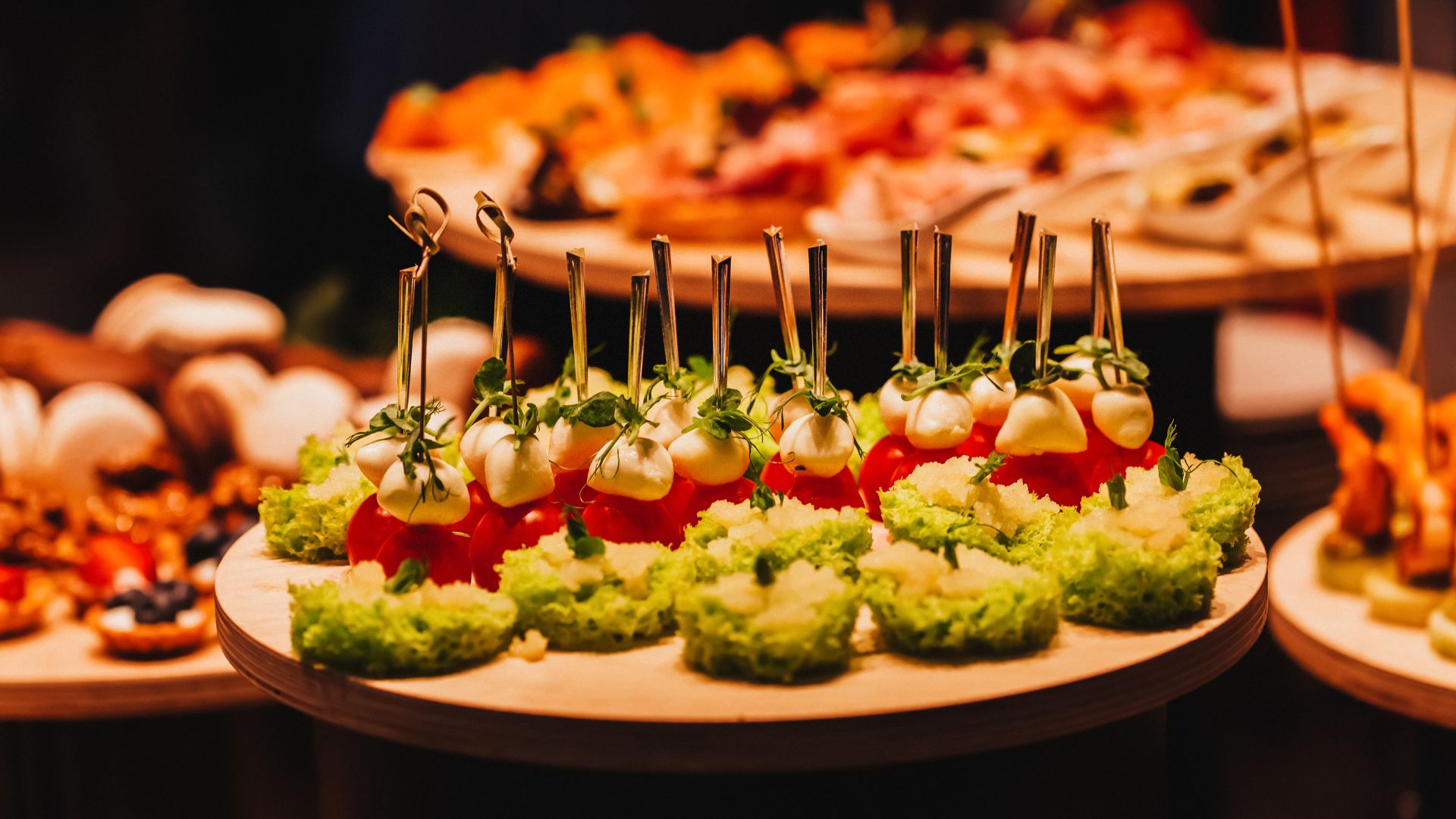 Close-up of a catering platter with decorative finger food, featuring skewers of cherry tomatoes, mozzarella balls, and fresh greens, alongside bite-sized green canapés, arranged on a wooden serving stand with other appetizers blurred in the background.