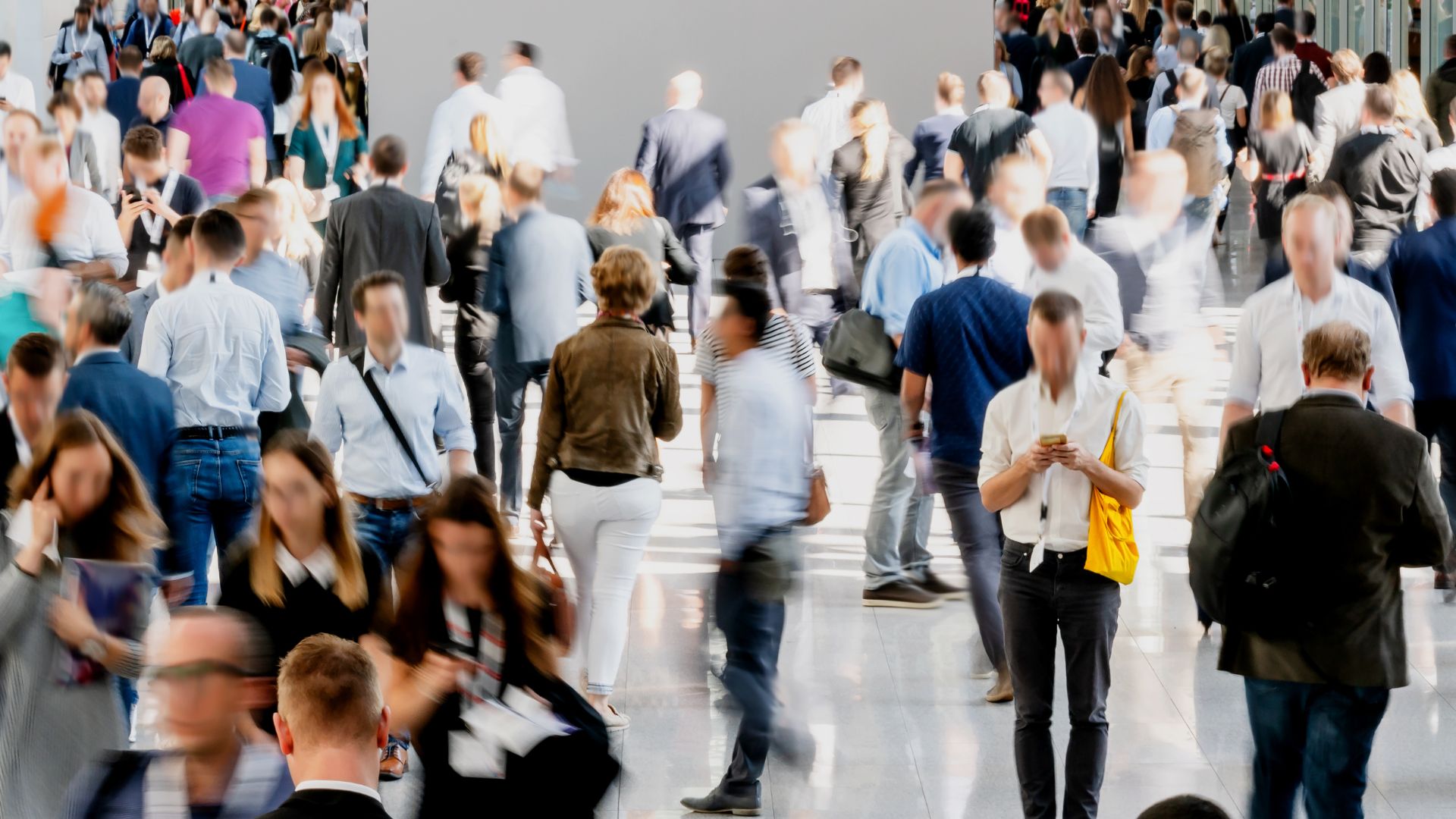 Blurred motion shot of a large group of people walking through a bright, spacious venue, many dressed in business attire, creating a dynamic scene of networking and movement.