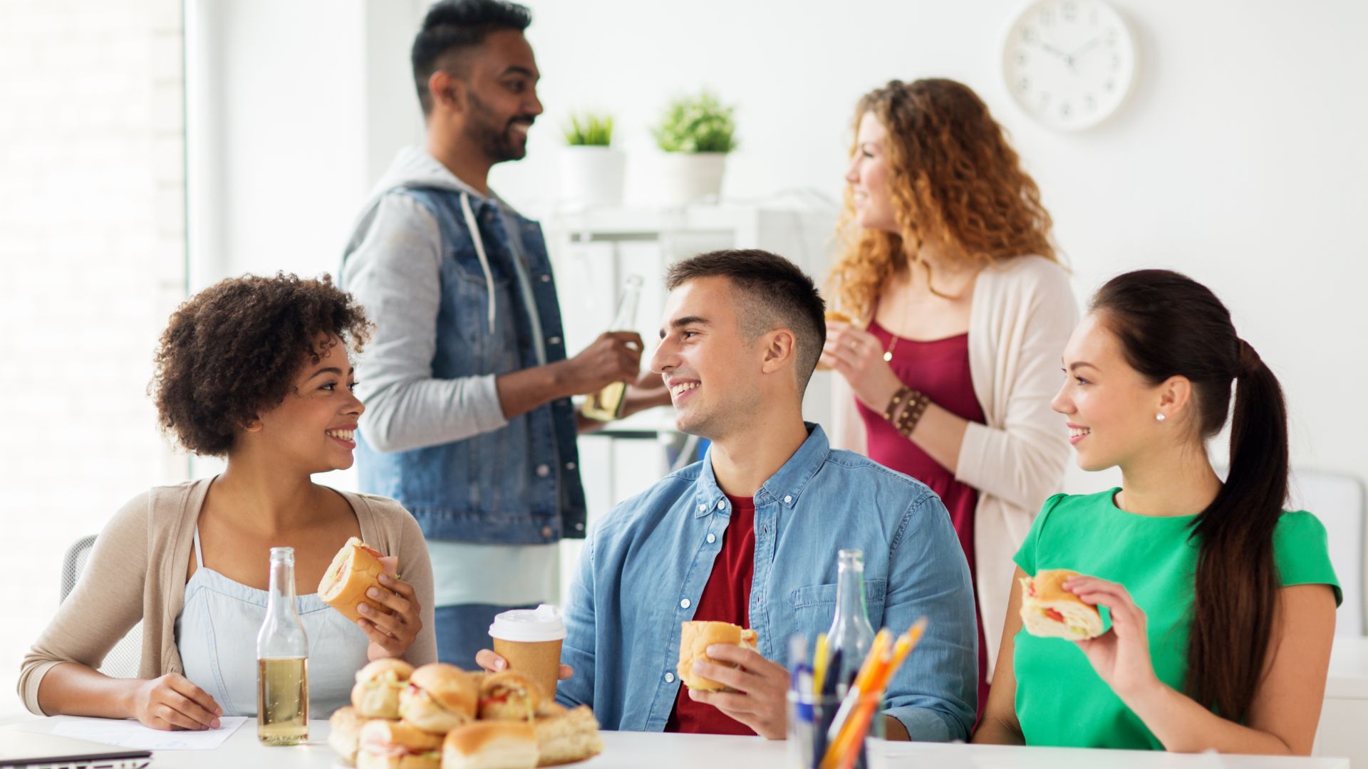 Colleagues enjoying sandwiches and drinks together during a relaxed office lunch break.