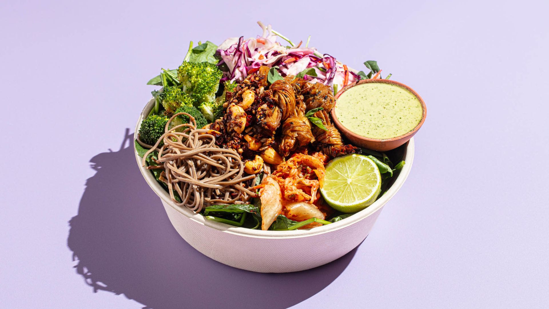 Colorful office catering bowl with soba noodles, broccoli, kimchi, lime, and green sauce, representing fresh, nutritious workplace meals