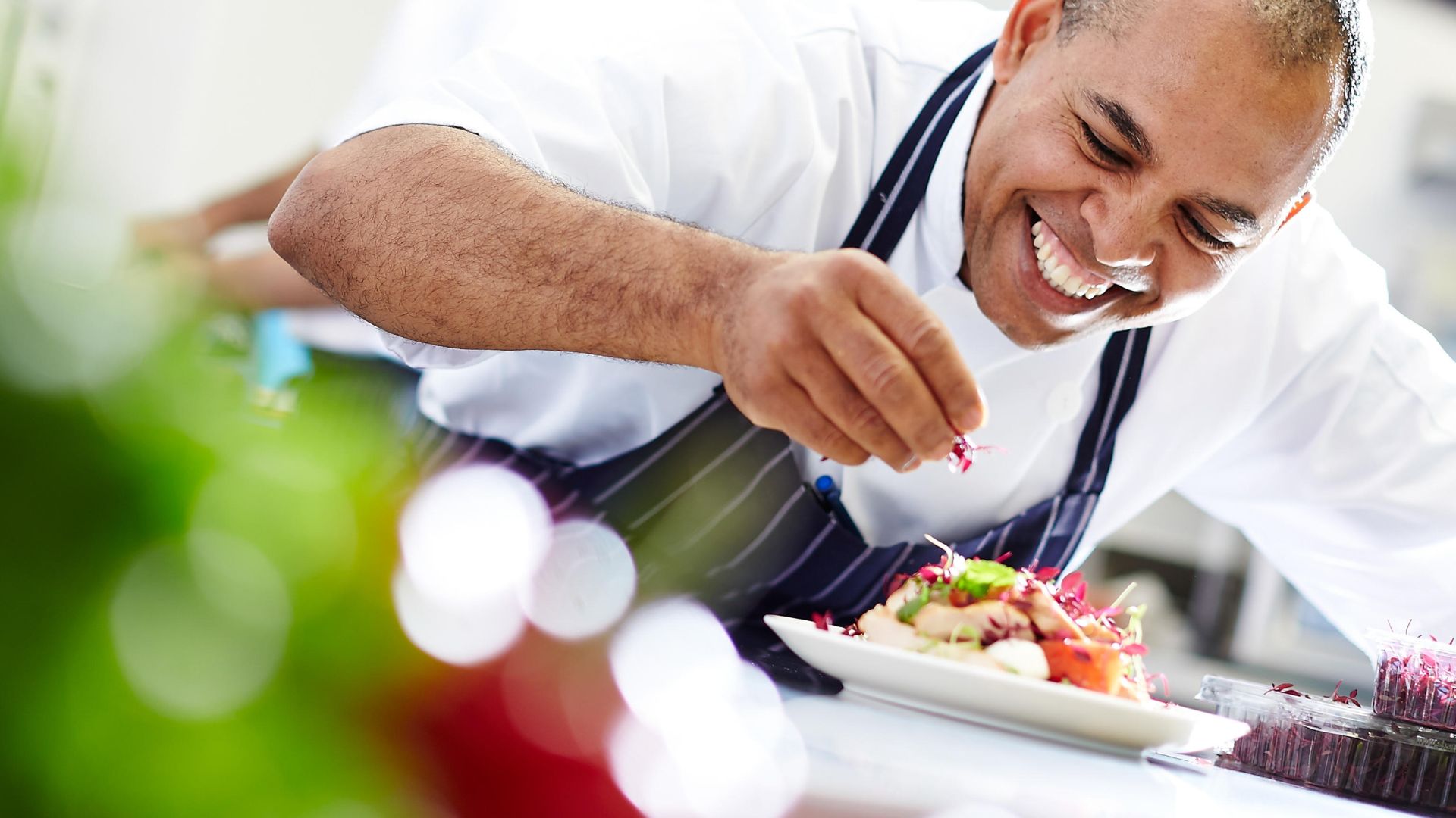 Smiling local chef adding garnish to a plated dish in a professional kitchen, highlighting handcrafted food and independent vendor quality