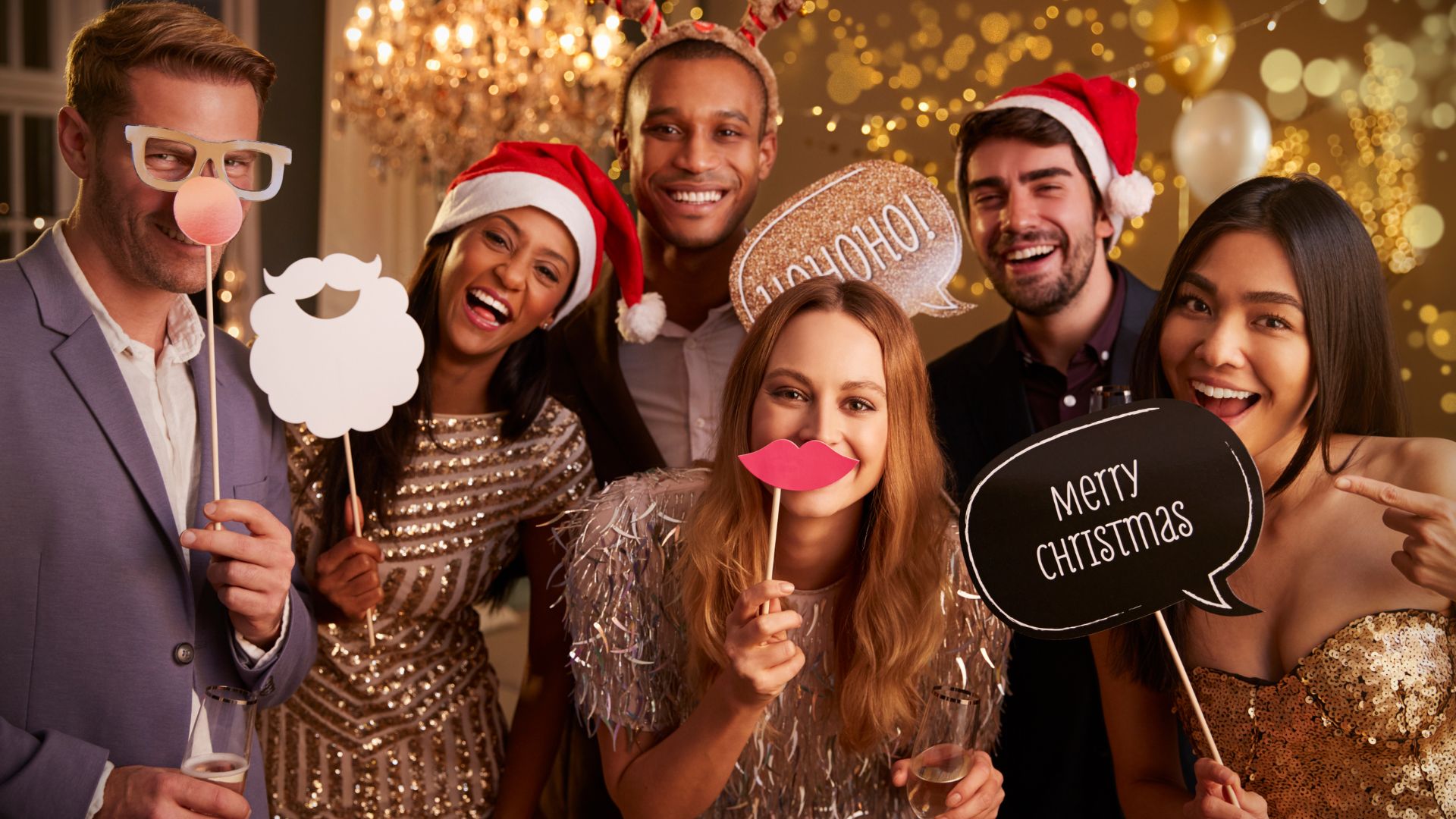 Group of cheerful coworkers at a festive office Christmas party wearing Santa hats and holiday props while posing for a photo booth picture.
