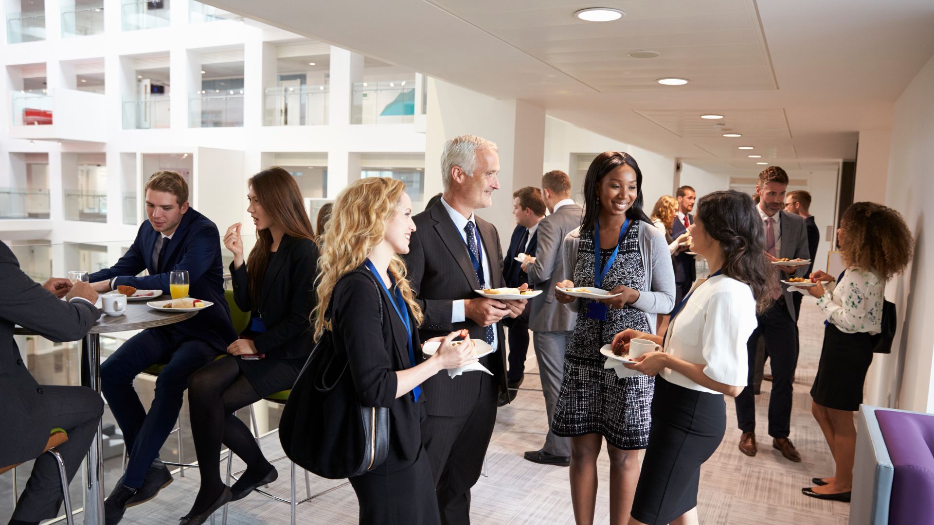 Business professionals networking during a catered office event, holding plates and coffee in a modern atrium.