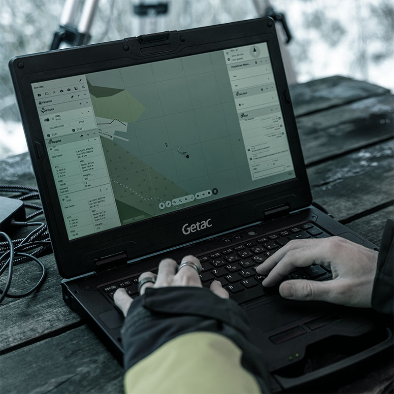 Hands typing on a Getac laptop displaying a navigation map interface on a wooden surface.