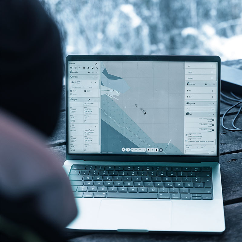 Laptop on a wooden surface displaying a navigation interface with maps, vessel and target data, and communication logs.
