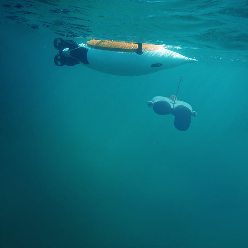 Underwater view of a white underwater drone with a yellow top and a separate remote control device floating in clear ocean water.