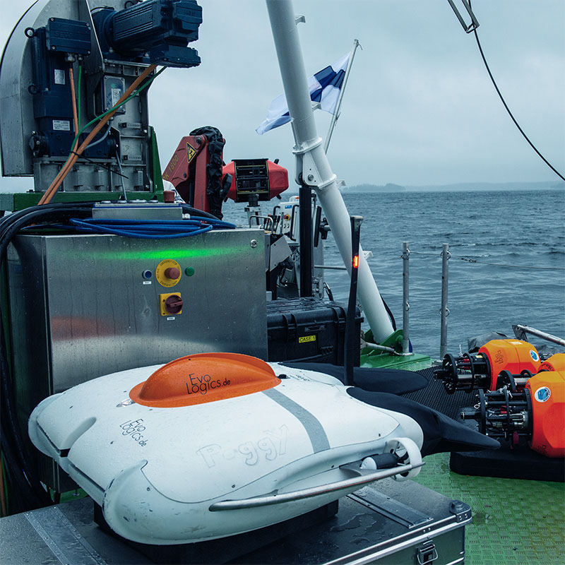 White autonomous underwater vehicle with orange top labeled 'Poggy' on a green deck near control equipment, with sea and cloudy sky in the background.