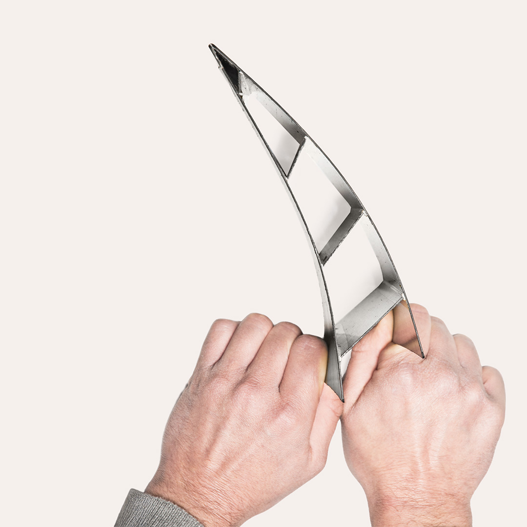 Two hands holding a curved metal fin structure with rectangular cutouts against a white background.