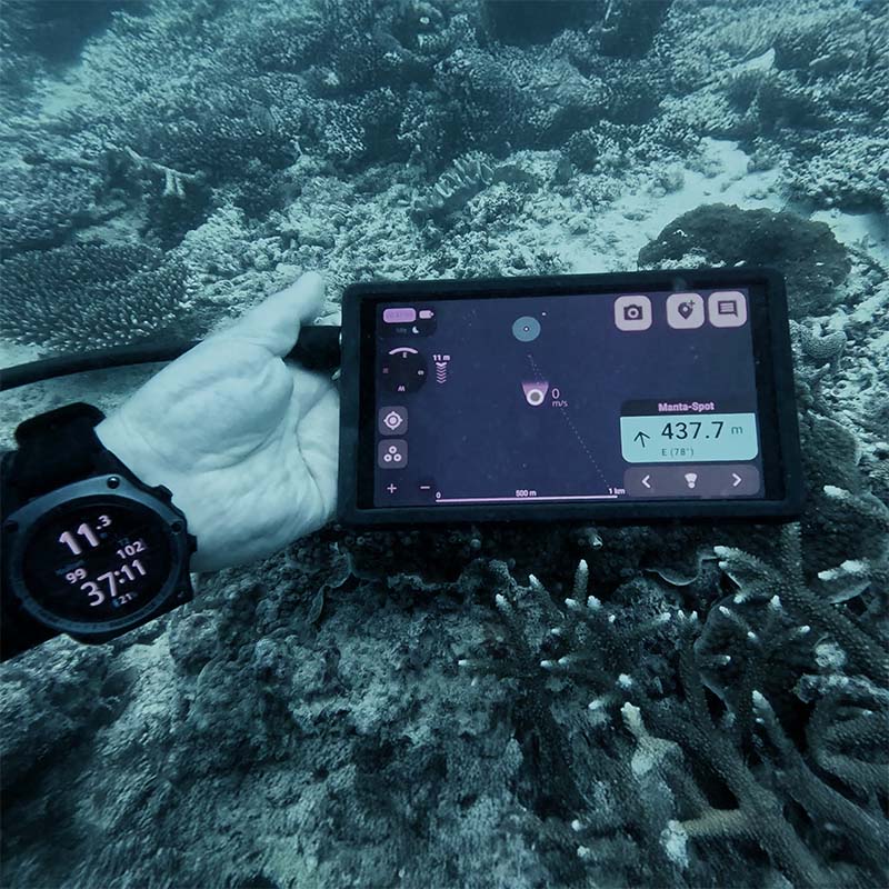 Diver's hand wearing a dive computer wristwatch holding an underwater navigation device displaying distance and location data over coral seabed.