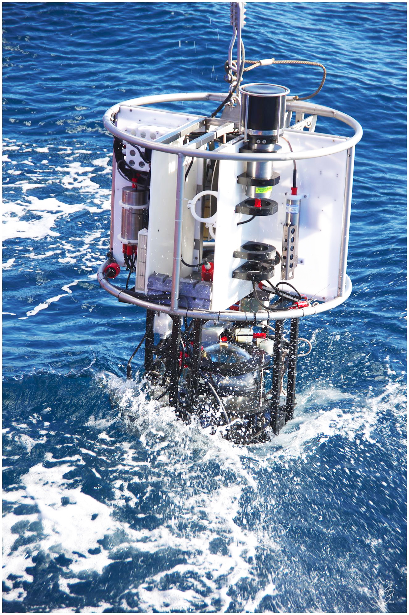 Oceanographic instrument being lowered into blue sea water with waves and splashes.