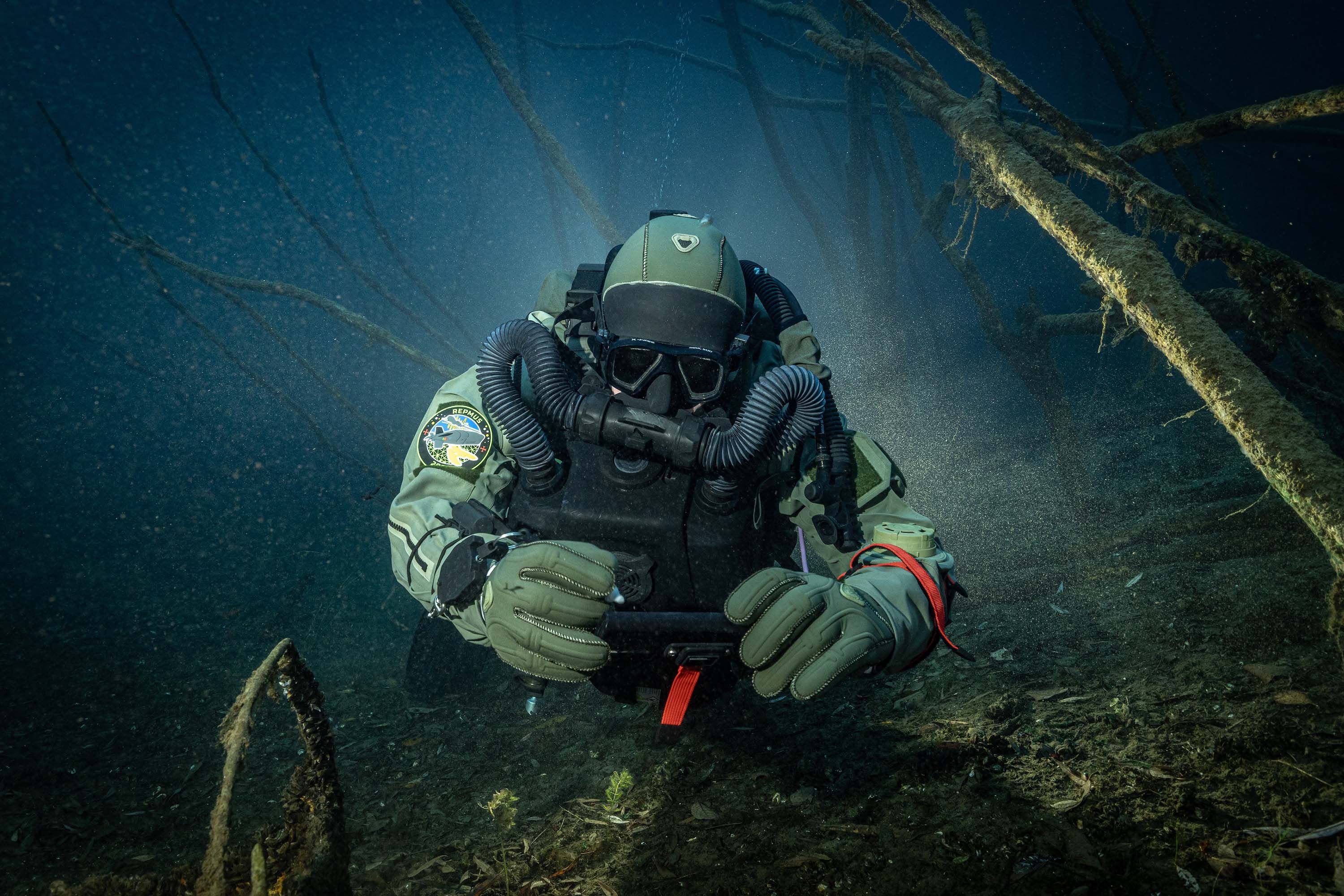 Scuba diver in specialized gear swimming underwater near submerged tree branches.