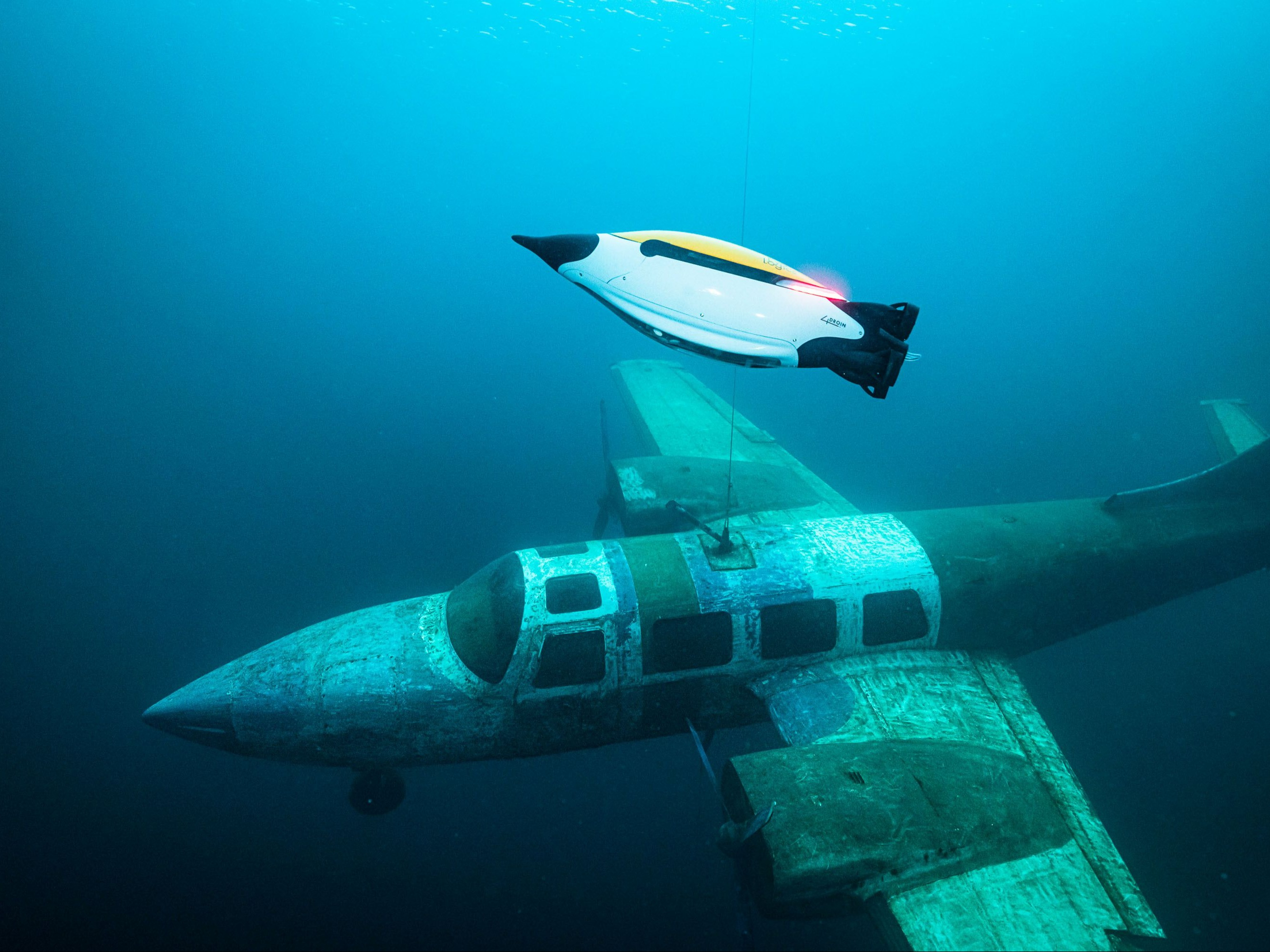 Underwater drone hovering above a sunken small airplane in a deep blue ocean.