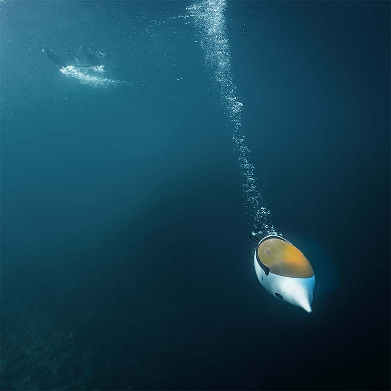 White underwater vehicle with a transparent orange dome descending into deep blue water, trailing bubbles toward the surface.