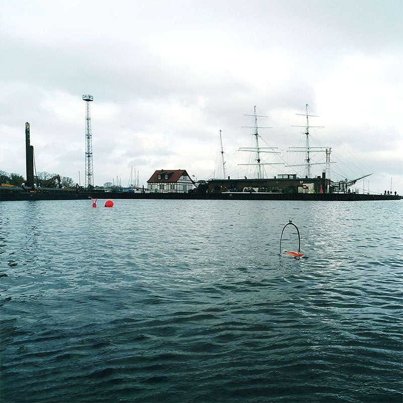 Harbor scene with a historic tall ship docked near buildings and industrial structures under a cloudy sky.