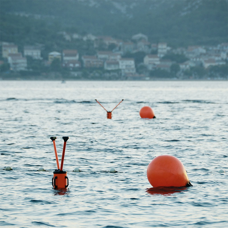 Orange buoys floating on calm water with a blurred shoreline of houses and hills in the background.