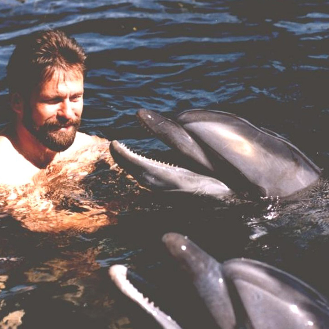 Bearded man smiling while swimming with two dolphins in dark water.