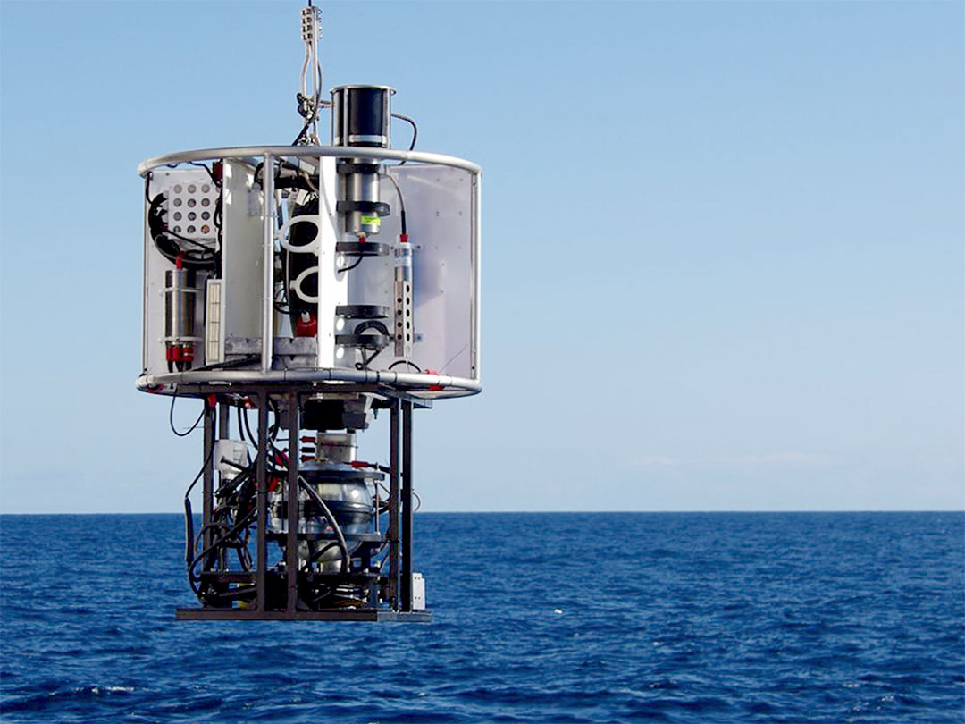 Marine scientific instrument suspended above ocean water under a clear blue sky.