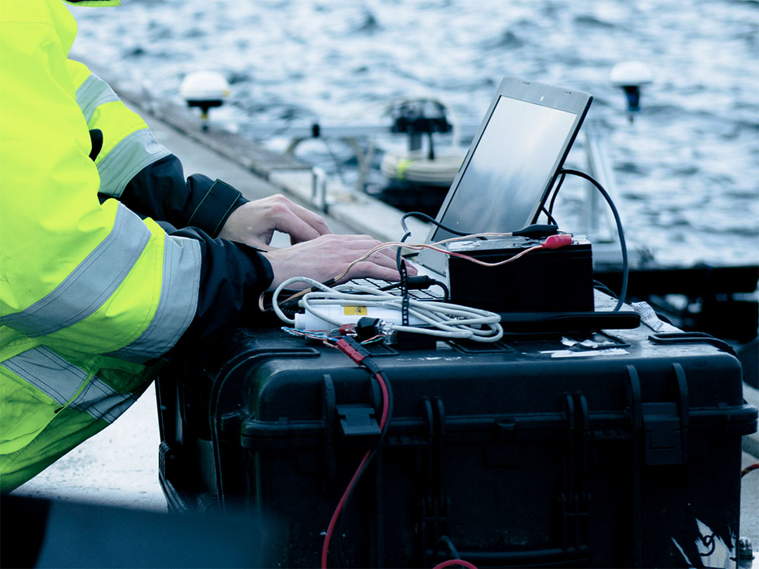 Person in high-visibility jacket working on a laptop connected to electronic equipment near water.
