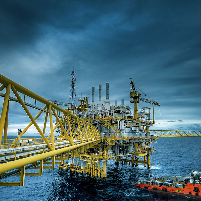 Yellow offshore oil rig platform with cranes and pipes extending over blue ocean under cloudy sky, with an orange supply boat nearby.