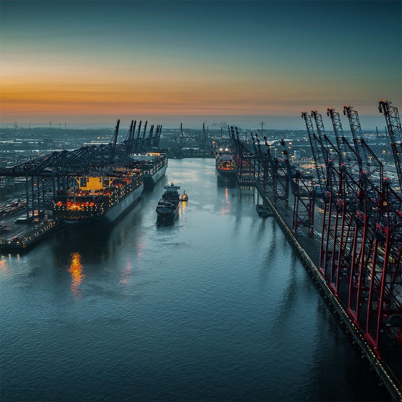 Evening view of a busy port with container ships docked and tall cranes along the waterway under a colorful sunset sky.
